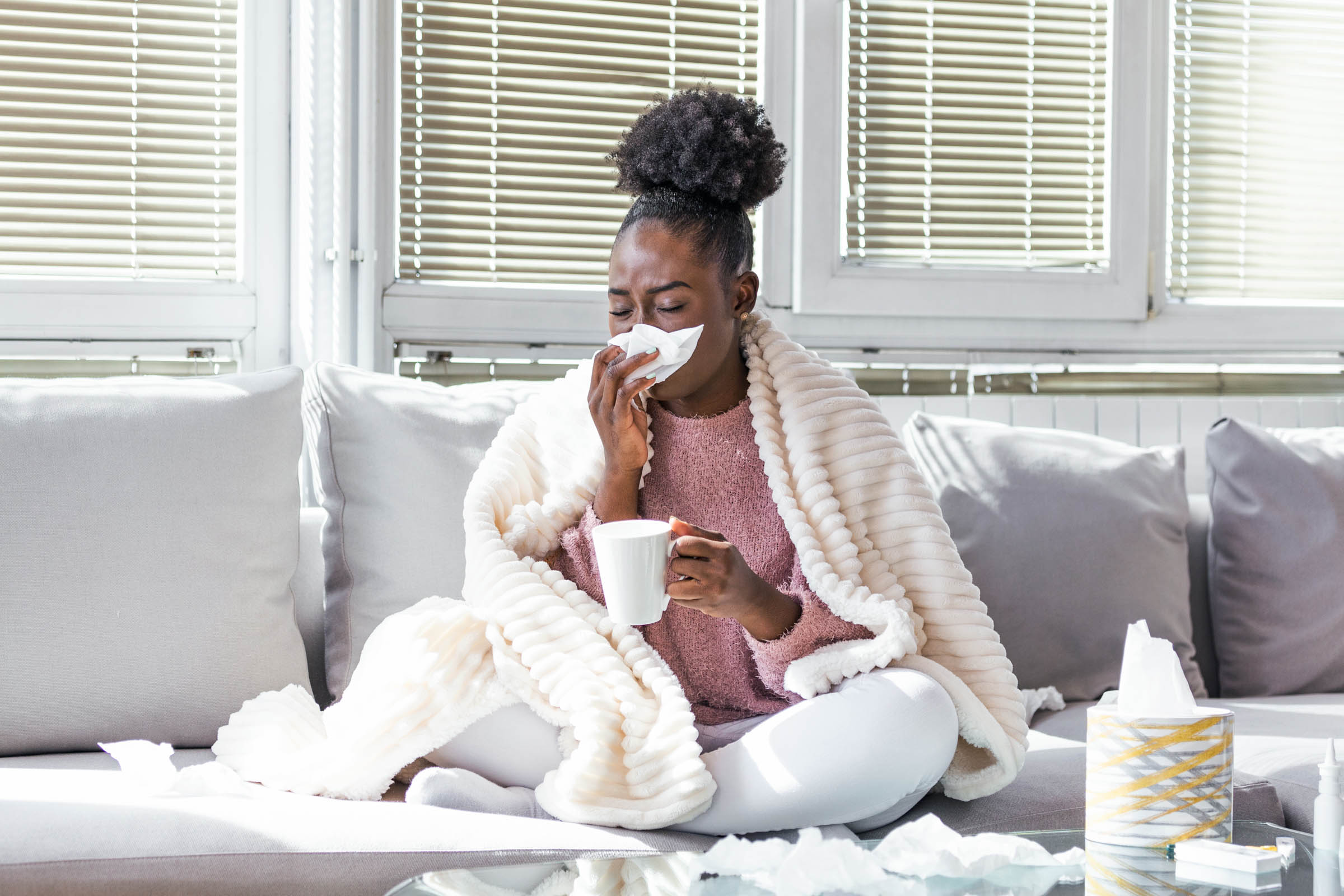Woman wrapped in a blanket sneezing into a tissue while holding a mug on a couch, suggesting flu recovery at home.