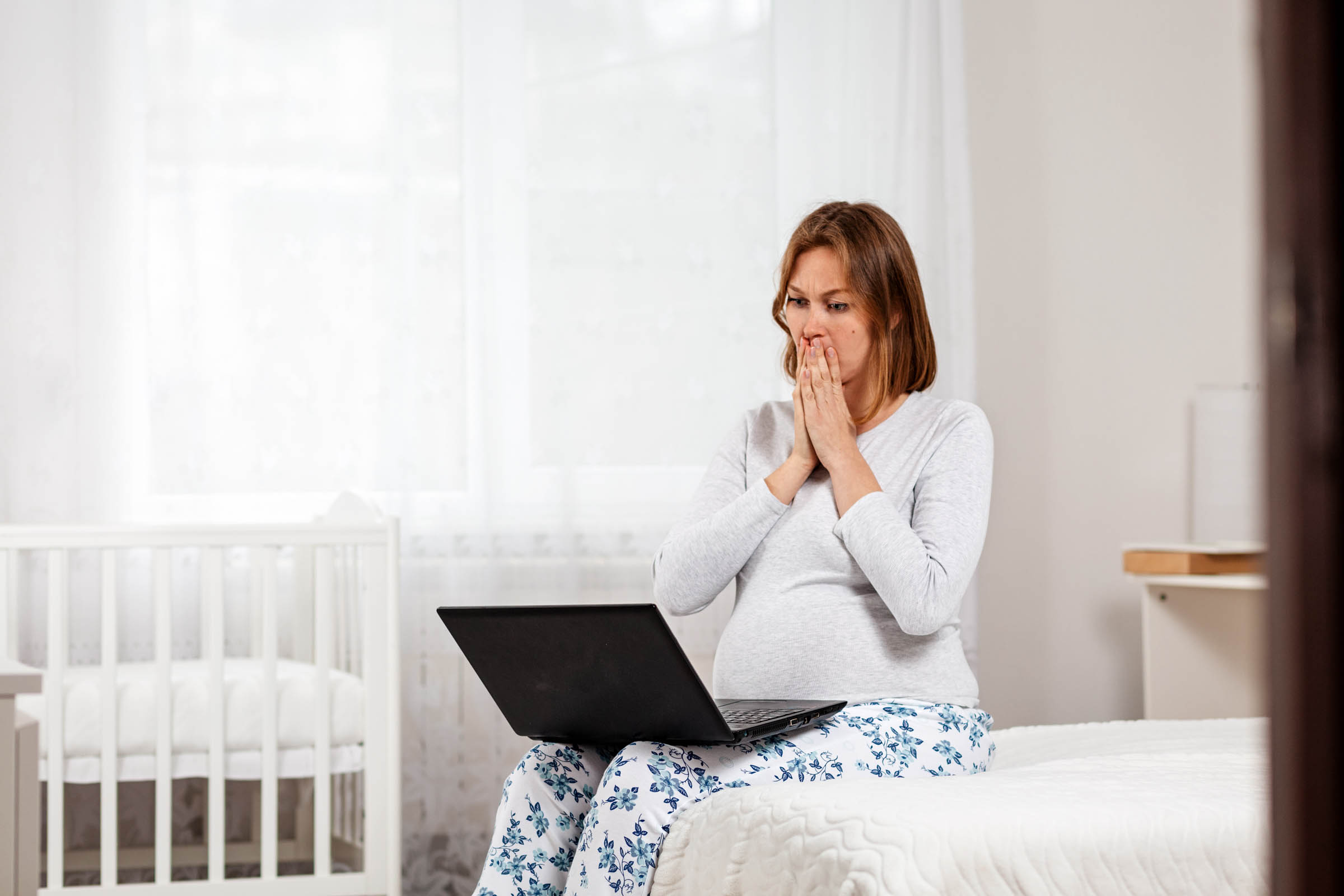 A pregnant woman sitting on a bed in a well-lit room, looking at her laptop screen with concern, her hands covering her mouth, with a crib in the background.