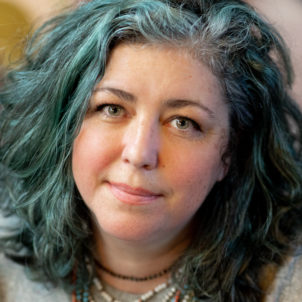 A portrait of Kimberly Sevilla, CEO of Shelter Air, with teal-gray wavy hair, a warm smile, and wearing a gray sweater.