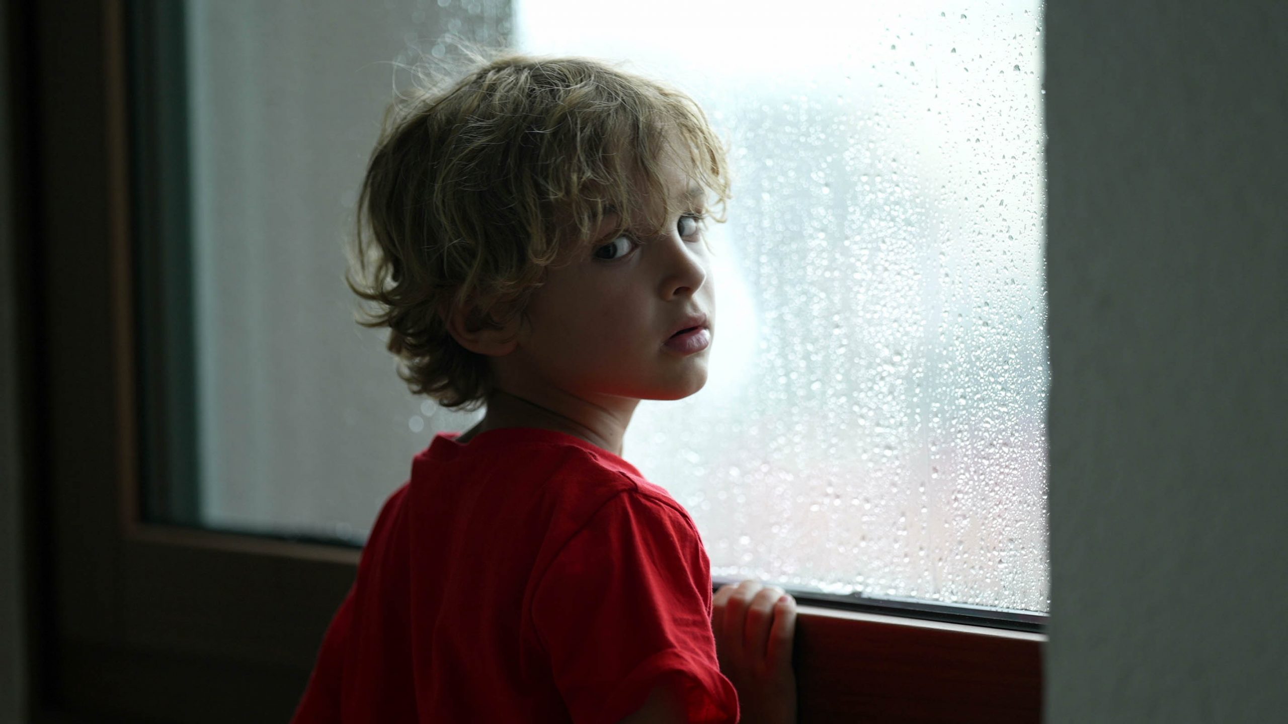 A thoughtful child in a red shirt looks out of a window dotted with raindrops, suggesting introspection and the quiet stillness of being indoors during rainy weather.