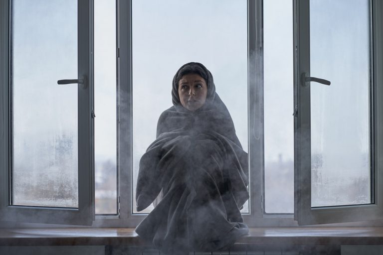 Person wrapped in blanket sitting by open window in cold winter weather showing need for ERV energy recovery ventilator in Beacon NY home.